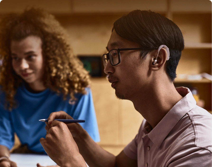 A person wearing glasses and an earpiece is holding a pen while sitting at a table with another individual in a classroom setting.